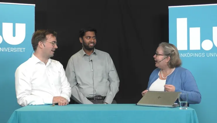 Three people stands in front of at a table with two blue background banners with the text ‘Linköping University’. They are talking and smiling at each other. A laptop and a glass of water are on the table.