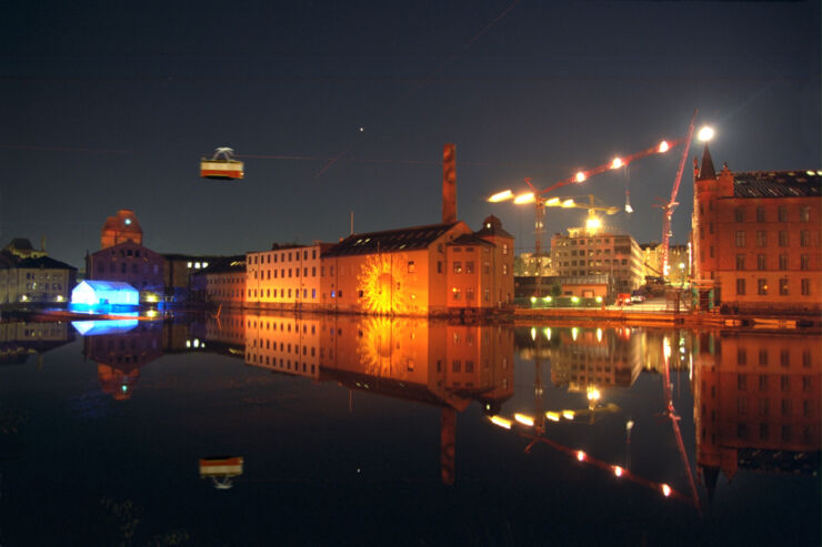 Illuminated buildings by a river at night. A tram flies above.