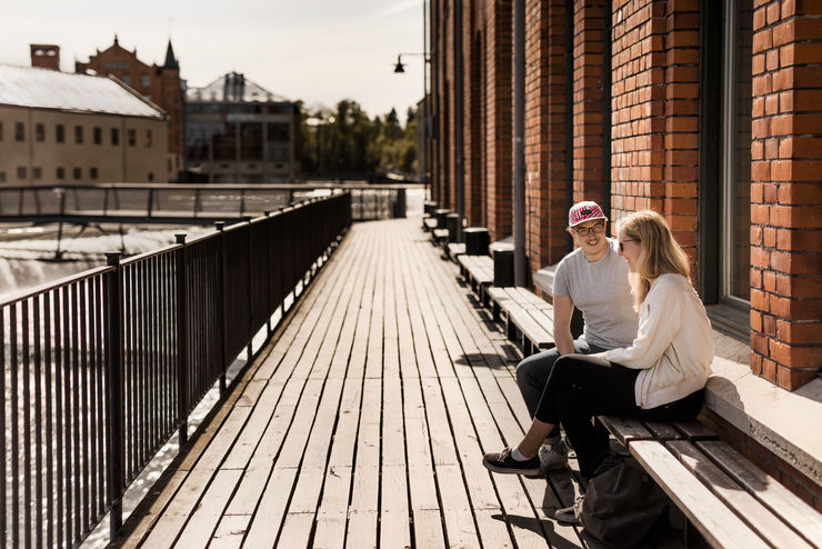 Två glada studenter sitter på en bänk vid strömmen genom Campus Norrköping