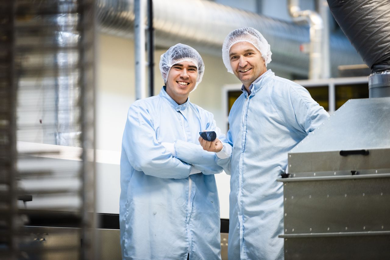 Two men wearing lab coats and hair nets smiles to the camera