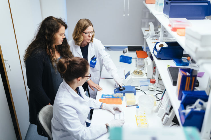 Three women, two of them wearing white lab coats, in front of a desk