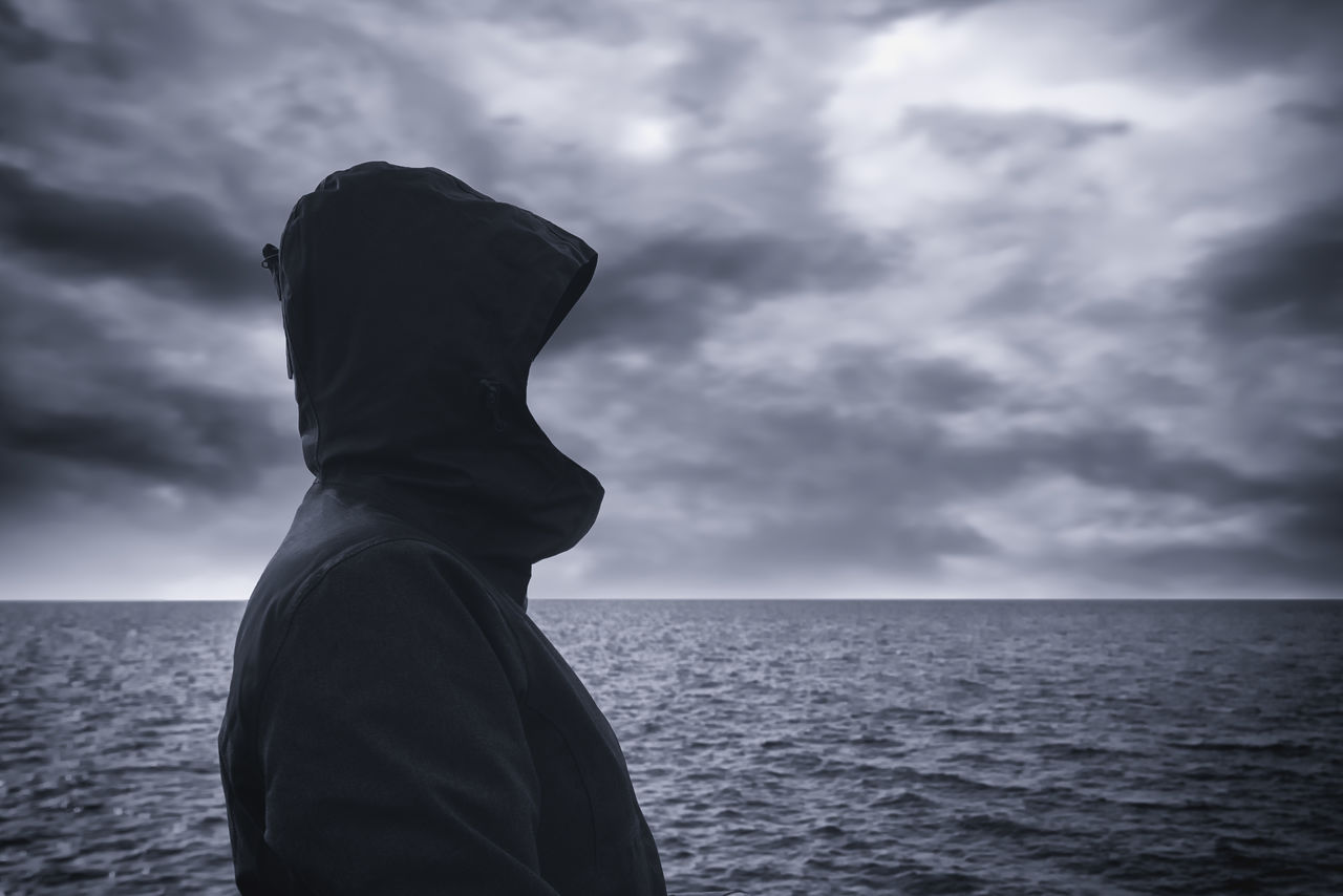 Faceless hooded person looking into distance at horizon over the sea water on cold windy winter day, anticipating unpredictable future