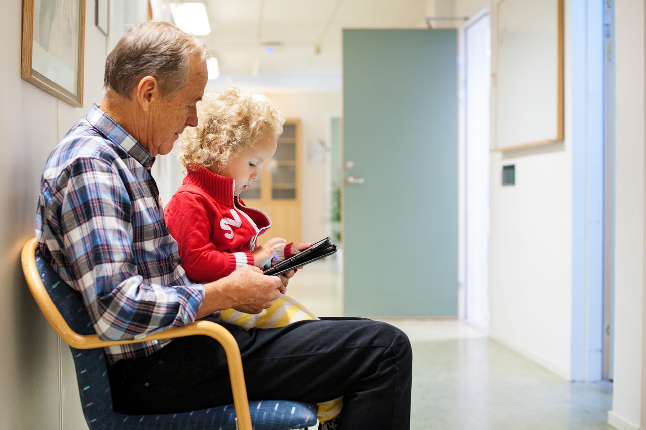 An elderly man and a small child are looking at a cell phone together. They are sitting on a couch in a hospital waiting room. 