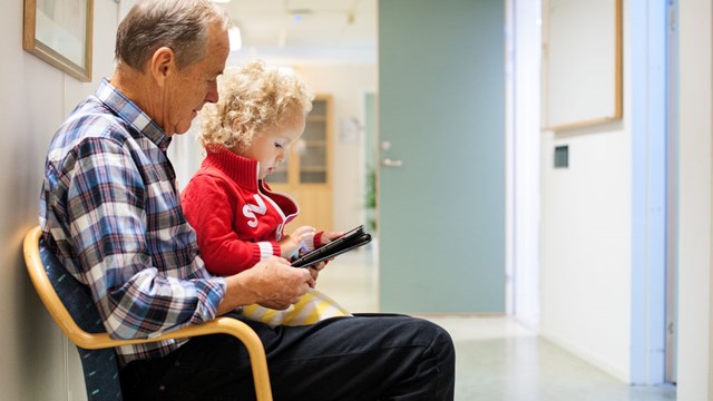 An elderly man and a small child are looking at a cell phone together. They are sitting on a couch in a hospital waiting room. 
