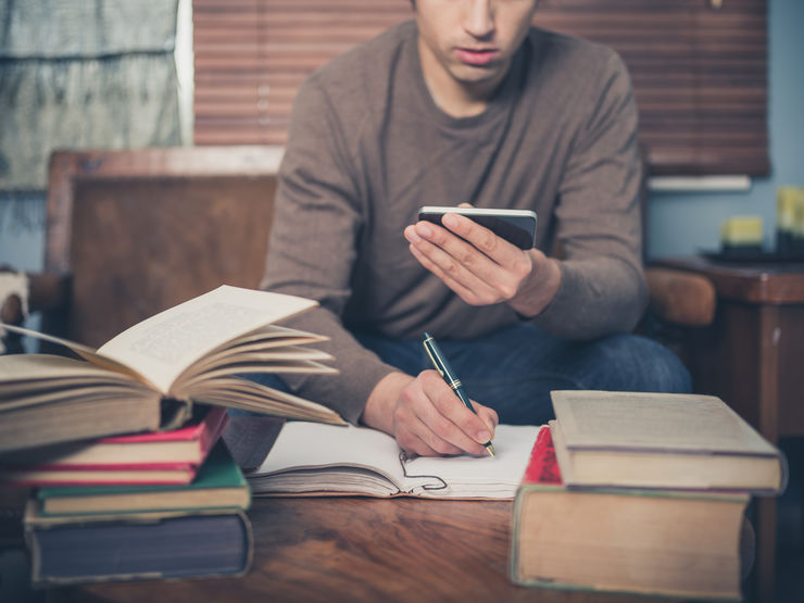 Young man sitting on couch with a cell phone in his hand. Books on the table in front of him