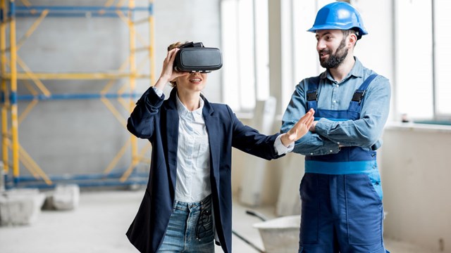 En kvinna, som bär VR glasögon, står bredvid en man med säkerhetshjälm på en byggarbetsplats.