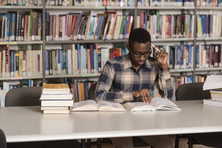 Portrait Of African Clever Student With Open Book Reading It In College Library - Shallow Depth Of Field