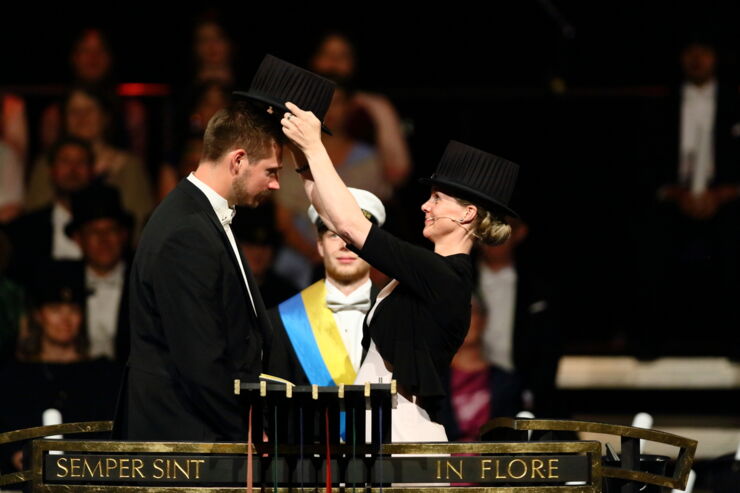 A woman wearing a tall black hat is putting a tall black hat on a mans head in a ceremony.