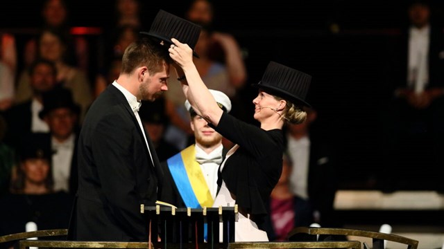 A woman wearing a tall black hat is putting a tall black hat on a mans head in a ceremony.