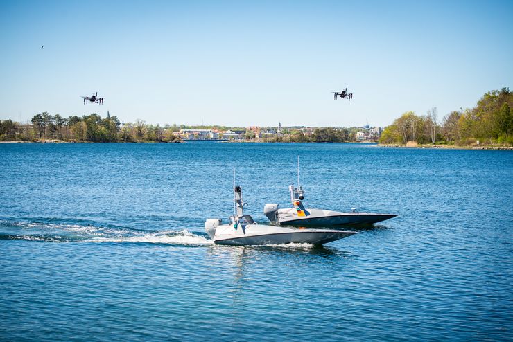 Två båtar på vattnet, i luften ovanför dem två drönare