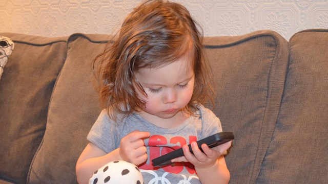 Toddler sitting on a couch, looking at a smartphone.