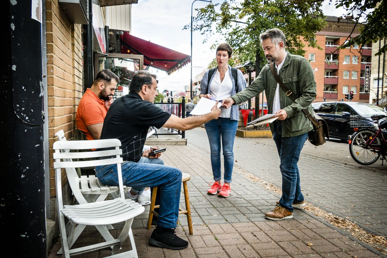 A man and a woman are handing out papers to two men sitting on chairs on the sidewalk next to a building.