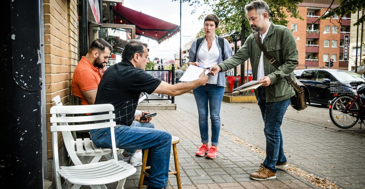 A man and a woman are handing out papers to two men sitting on chairs on the sidewalk next to a building.
