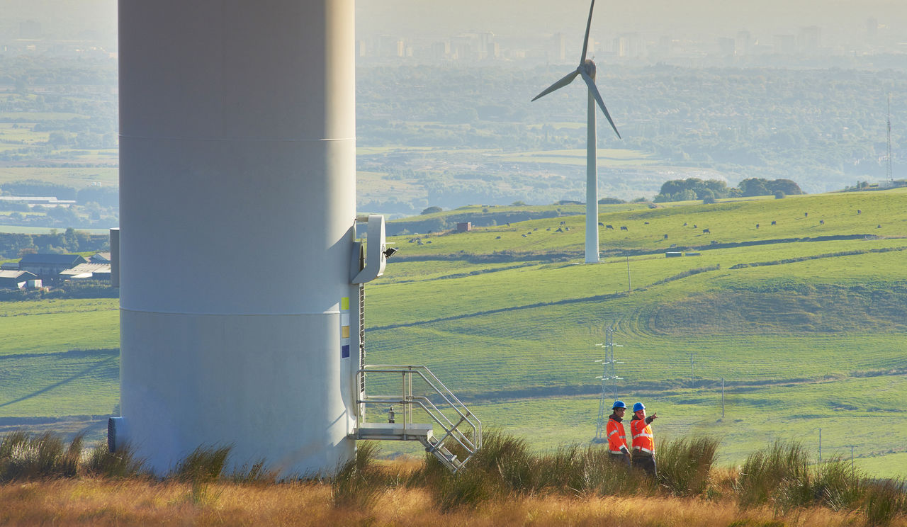 Two wind farm engineers walk away from the door way of a wind turbine. They are wearing orange hi vis jackets and blue hard hats. One is male , one is female. In the background wind turbines can be seen across the landscape down to a distant city.