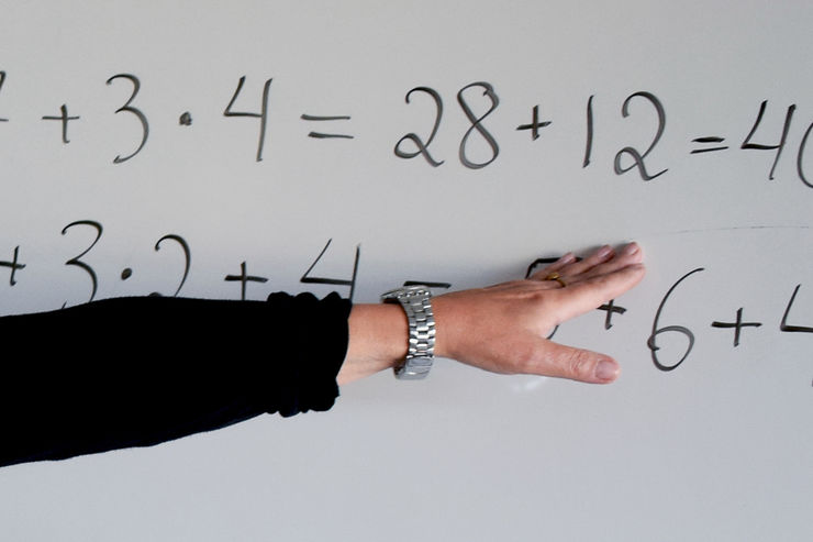 An arm wearing a black shirt and a watch is pointing to math problems written with black pen on a whiteboard 