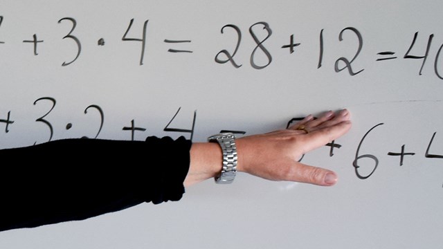 En klockbeprydd arm med svart skjorta pekar på mattetal skrivna med svart penna på en whiteboard