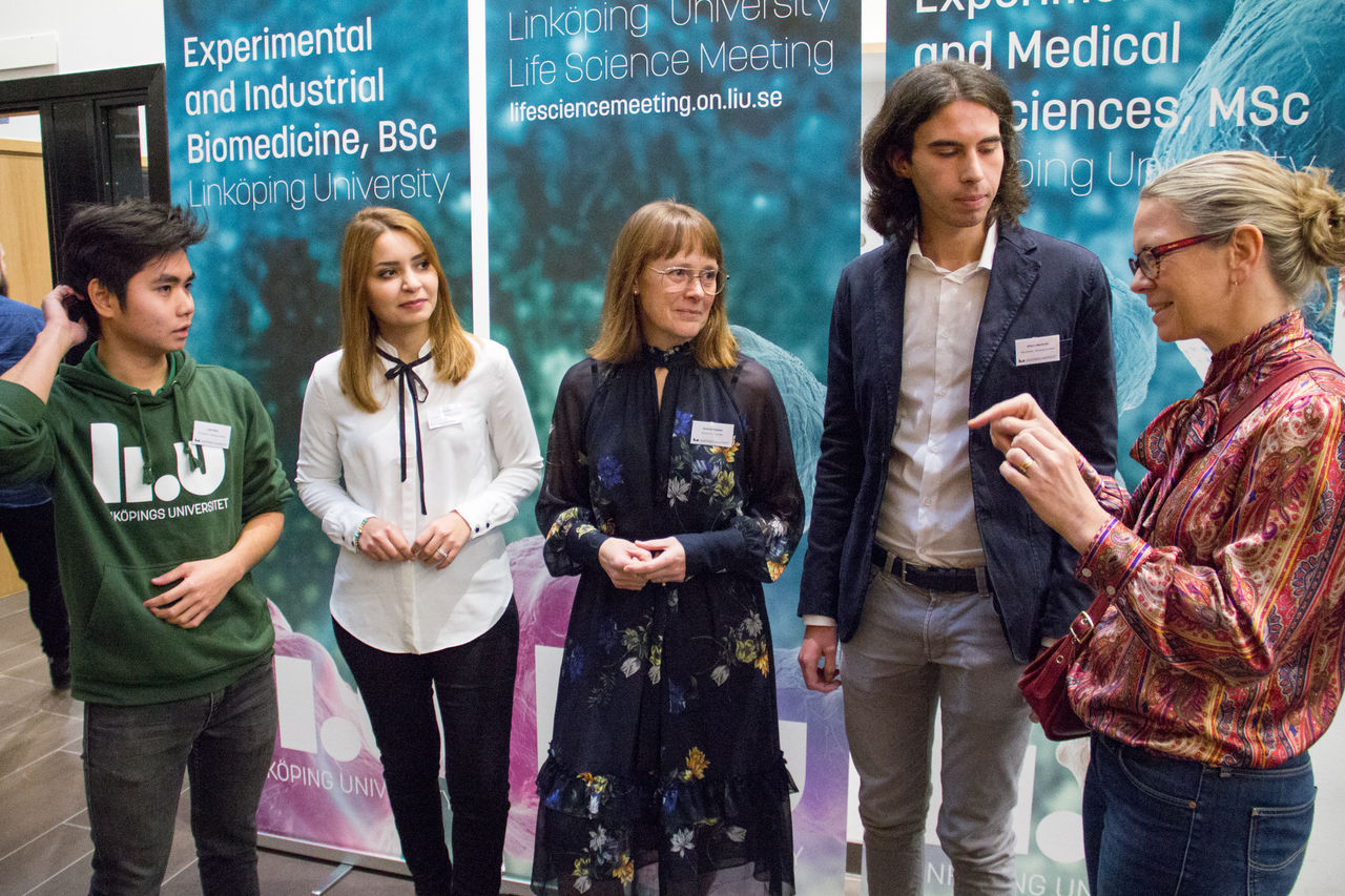 Fem personer samtalar framför en fotovägg.