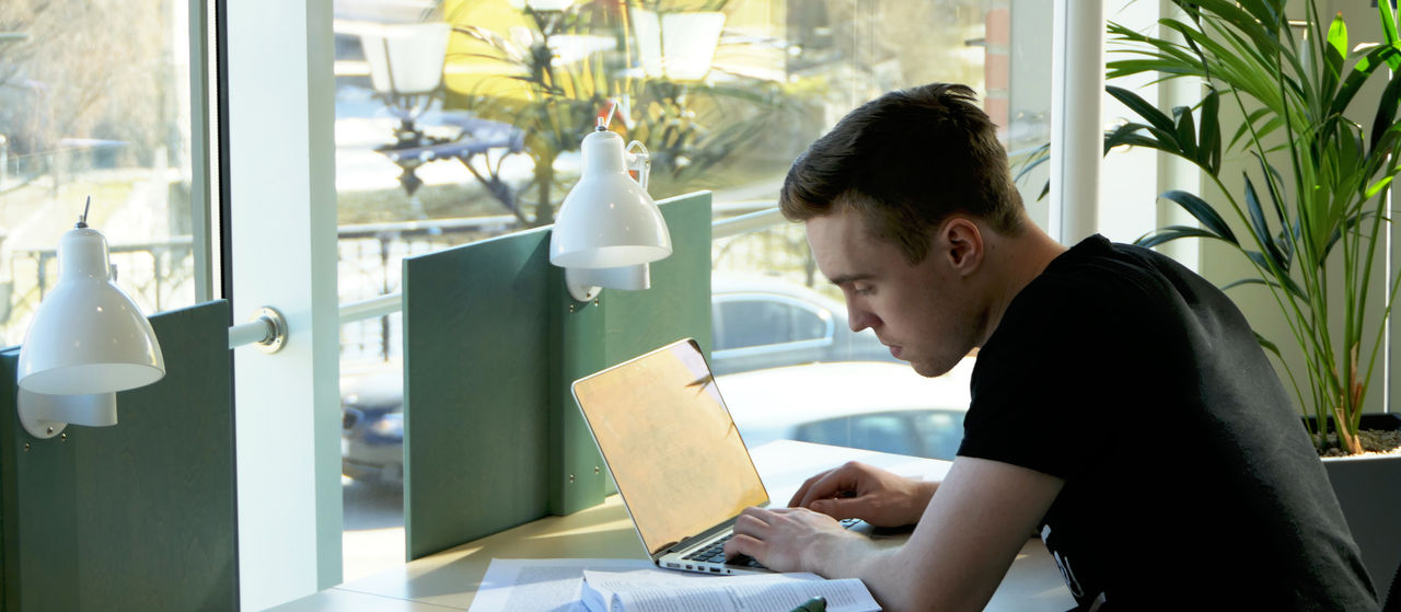 A male student with a laptop and a book by a window.