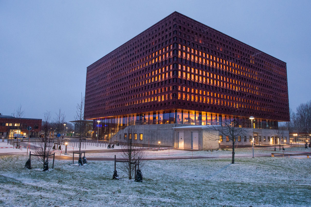 A big building in sunset, there is warm light coming from the windows, on the ground outside there is a bit of snow.