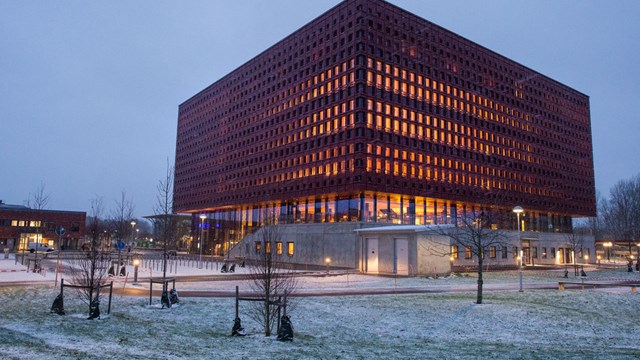 A big building in sunset, there is warm light coming from the windows, on the ground outside there is a bit of snow.