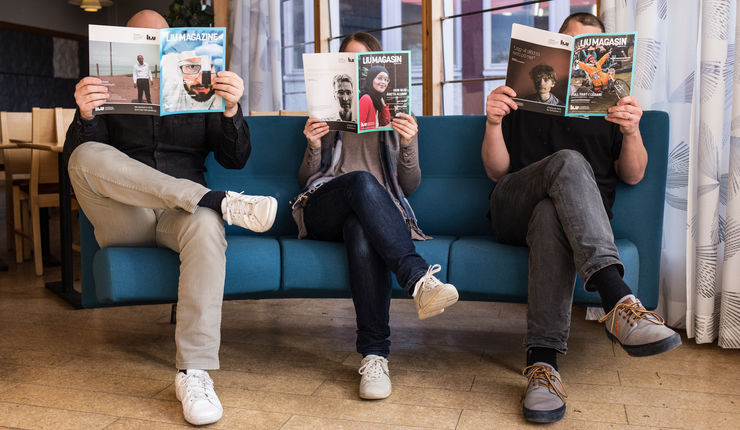 Three people sit in a sofa holding magazines so that they cover their faces