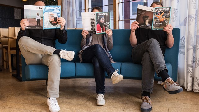 Three people sit in a sofa holding magazines so that they cover their faces