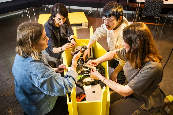 Four people kneeling around a yellow coffin filled with discarded cell phones and tablets. 