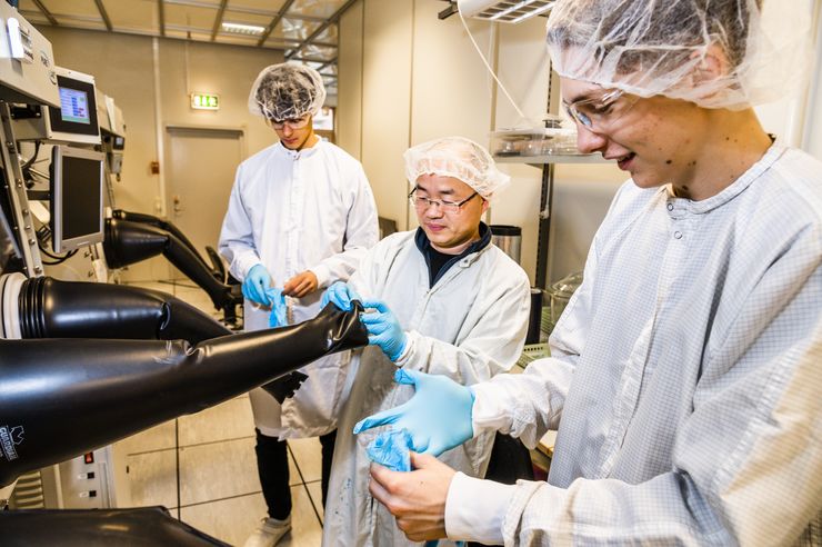 Tre personer i labbrockar, gummihandskar och hårnät står framför en stor maskin i labbmiljö.