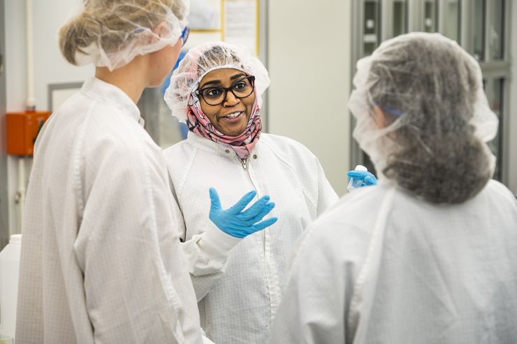 Two people with their backs to the camera are watching a woman who explains something to them. The picture is taken in a lab environment and everyone are wearing hair nets.