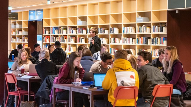 A number of students sitting a tables in a study space with a book shelf in the background.