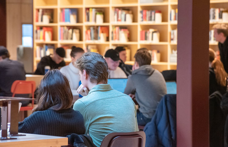 Students sitting at a number of tables with a book shelf behind them.