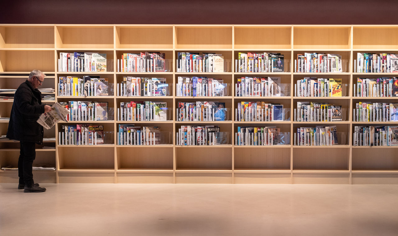 A shelf with magazines, and to the left a man standing and reading a news paper.