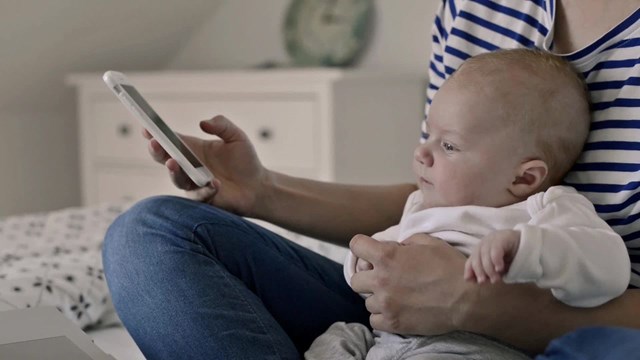 Unrecognizable mother with baby son, sitting on bed, holding a smart phone, texting, home office
