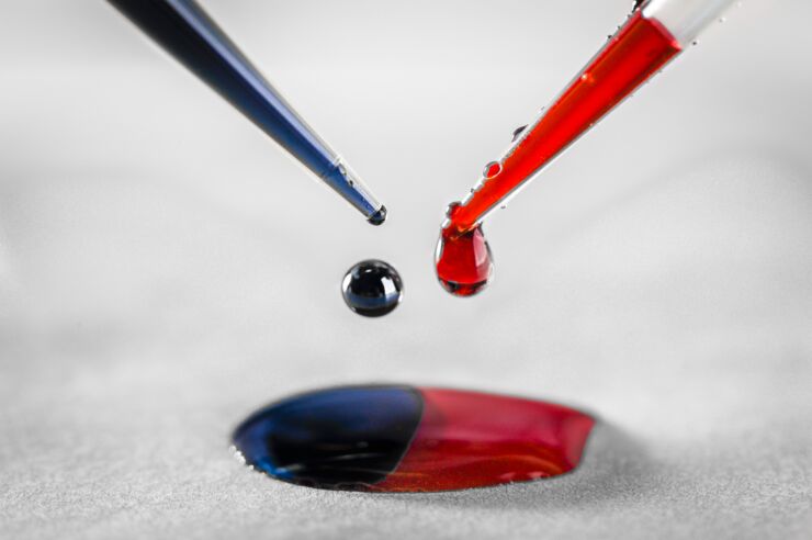 Closeup of two pipettes, one is dripping blue ink and the other one red ink. The ink is forming a two colored puddle.