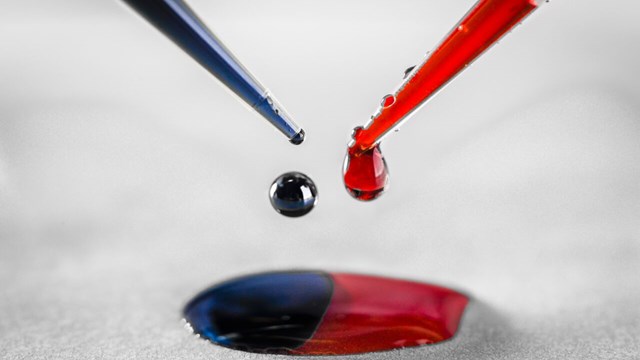 Closeup of two pipettes, one is dripping blue ink and the other one red ink. The ink is forming a two colored puddle.