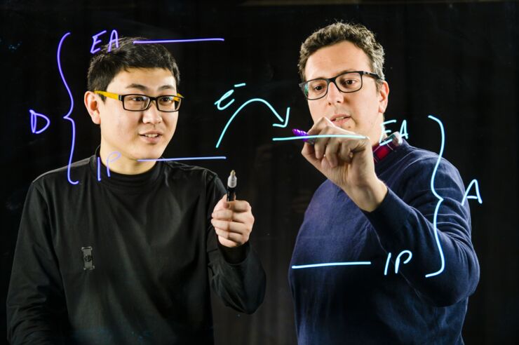 Kai Xu and Simone Fabiano photographed through a sheet of glass on which they are writing formulas.