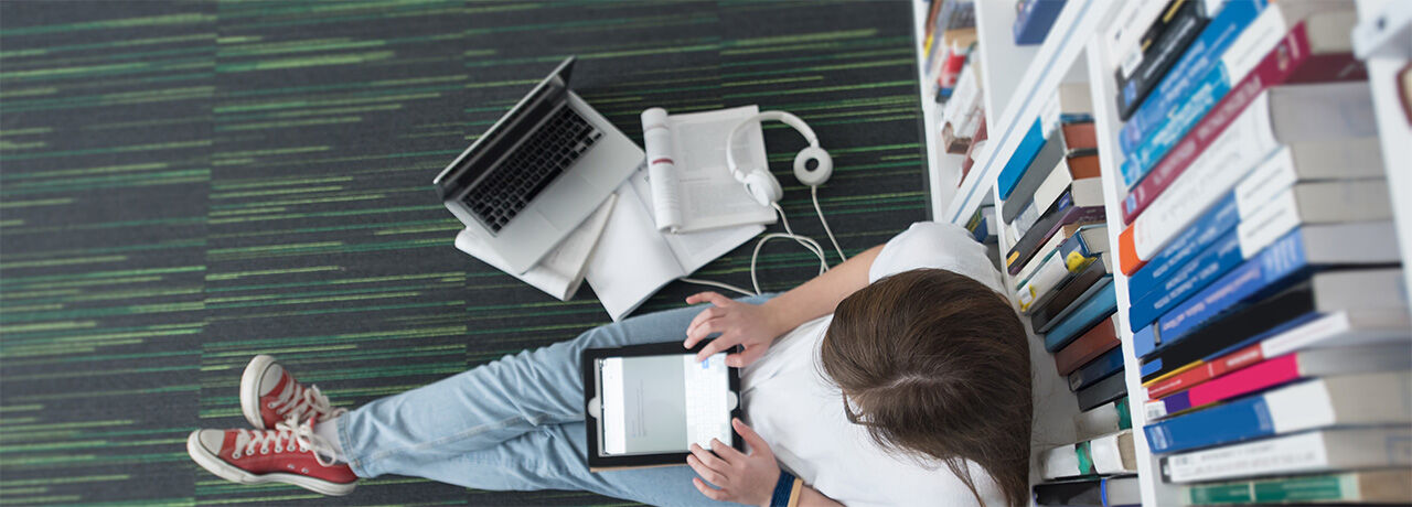 Kvinnlig student sitter på golvet, lutad mot bokhylla. Surfplatta i knät och laptop , hörlurar och böcker vid sidan om. Foto taget ovanifrån.