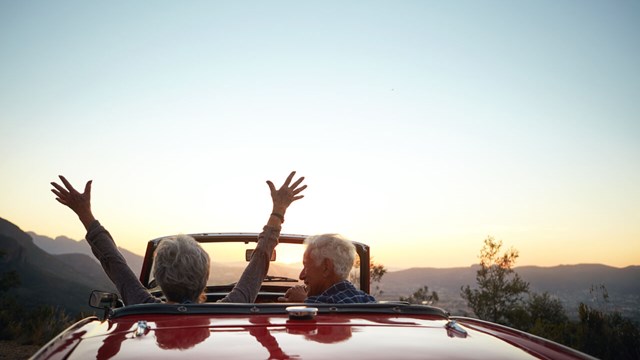 Older couple sitting in a car in front of a sunset. They have their backs to the camera. The woman sits behind the wheel and raises her hands in the air.