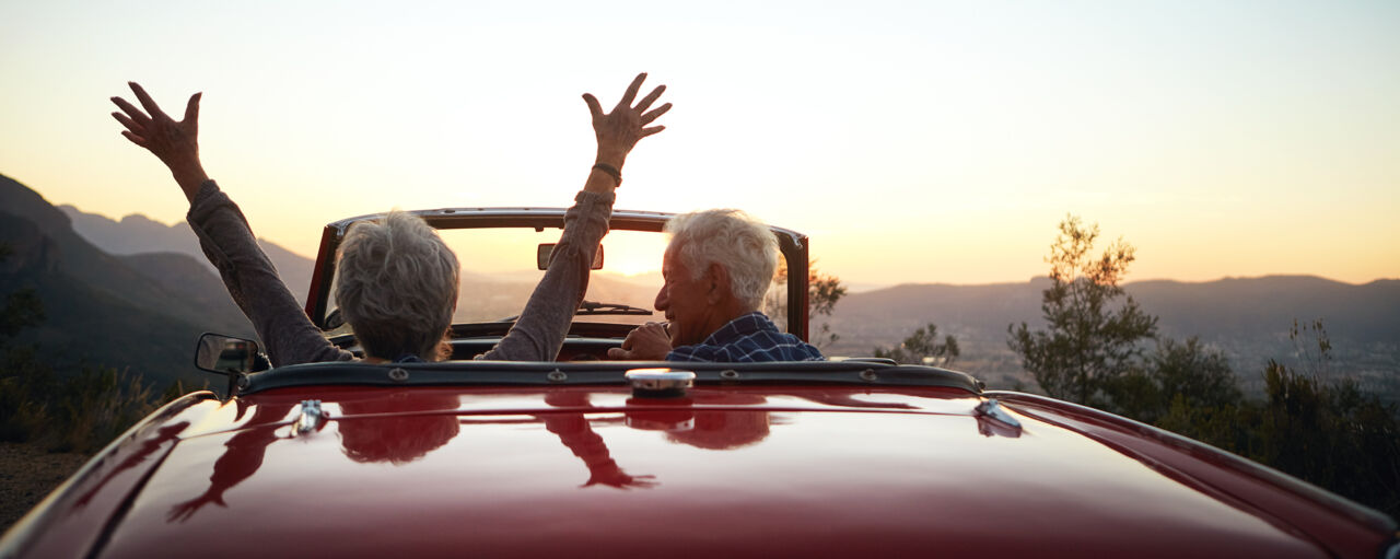An older couple sitting in a car at a view point and the woman, who is driving, is rasing her hands in the air. It's sunset.