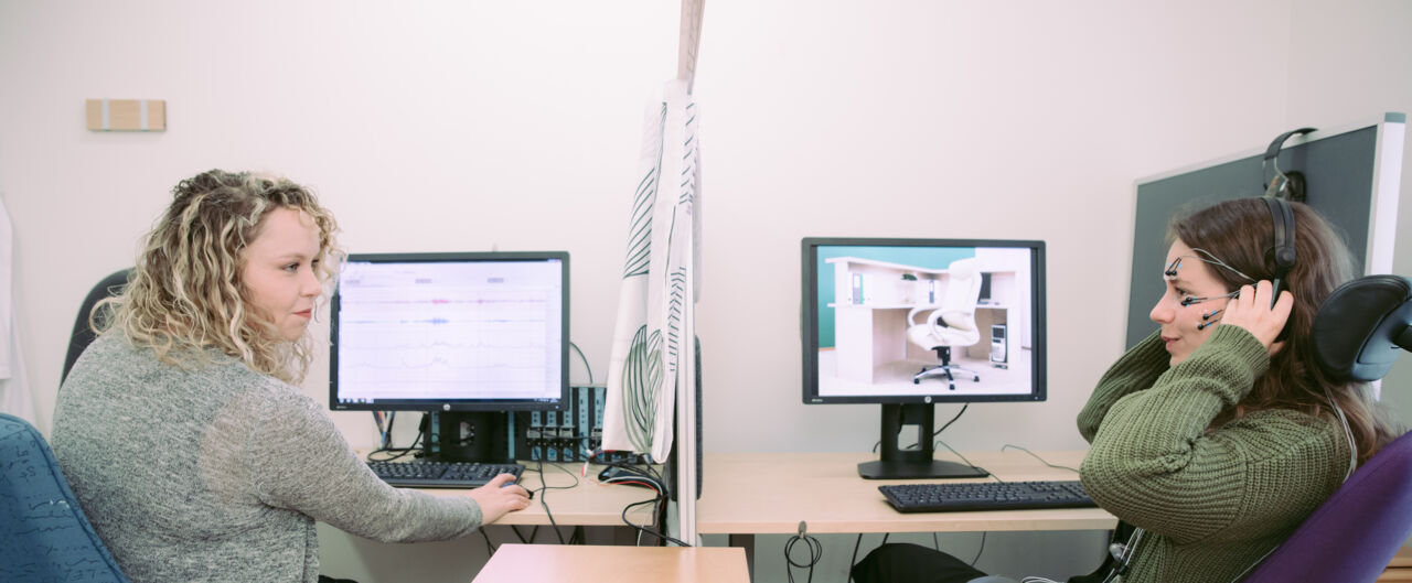 forskare och försöksdeltagare sitter vid varsin dator åtskilda av en gardin så att de inte ser varandra.