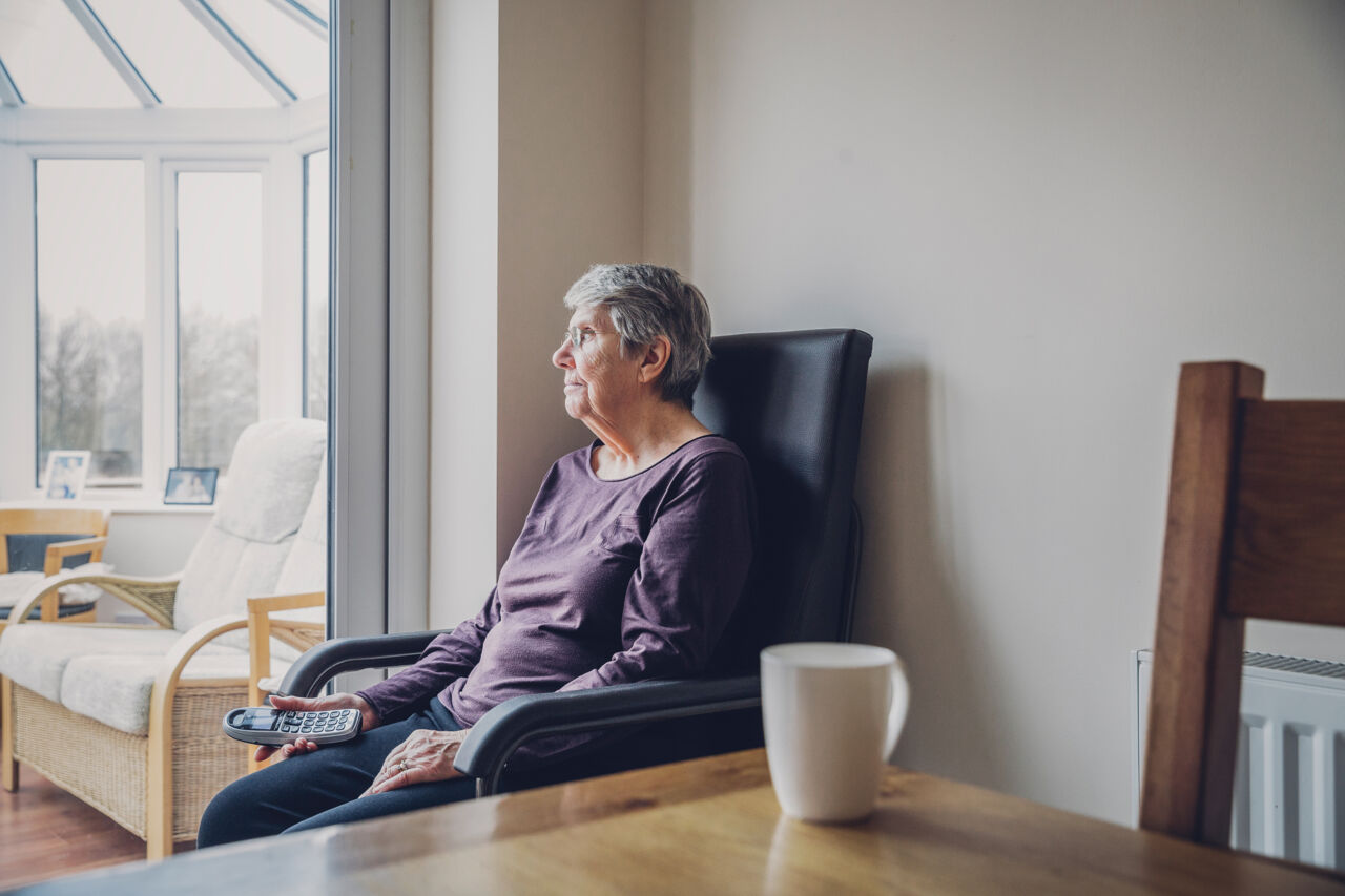 Senior woman sitting alone in her kitchen. She is looking out into her conservatory while holding a home telephone in her hand.