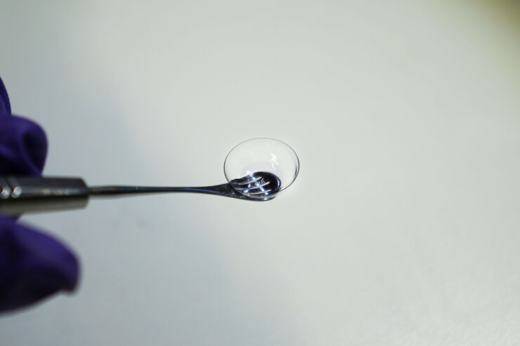 Close-up of a contact lens held up by a metal instrument. The background is gray.