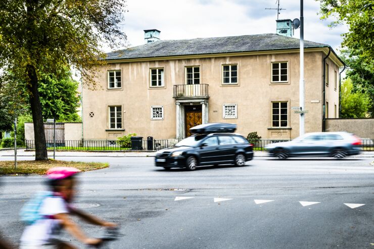 En stor beige villa. I förgrunden ett par bilar och ett barn på cykel i rörelseoskärpa.