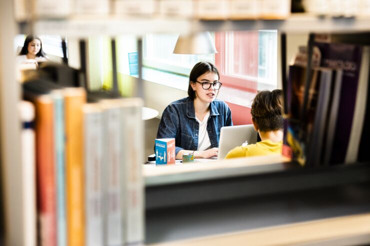 Three students seen in the space between rows of books in a book shelf.