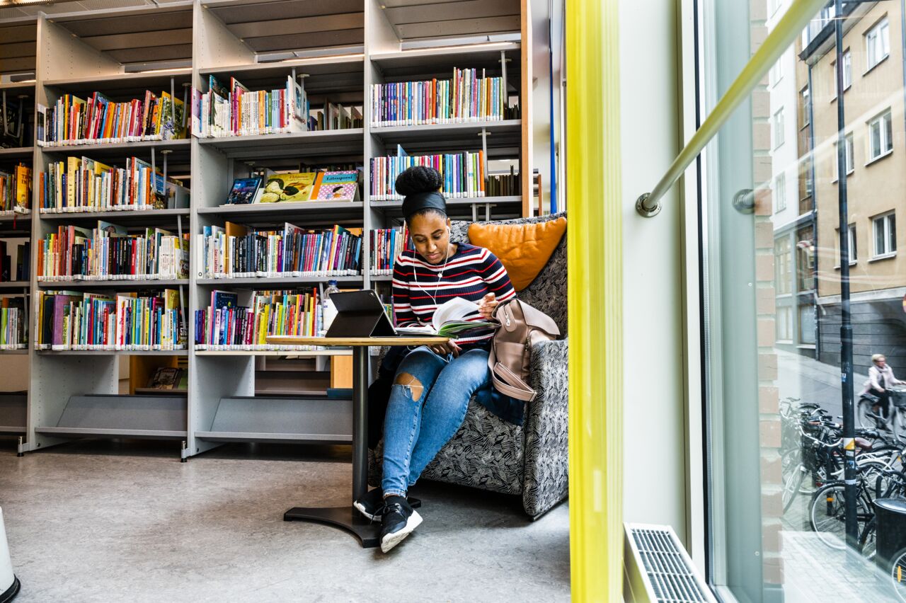 A female student seated in an armchair reading a book in front of a book shelf.