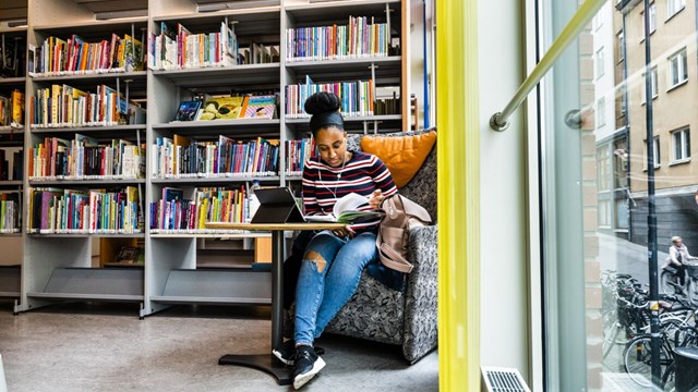En student som sitter i en fåtölj och läser en bok på ett bibliotek.