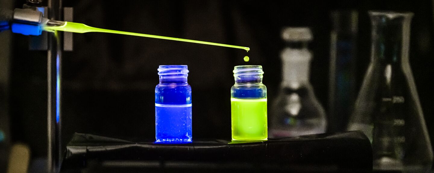 Two containers of blue and green fluorescent liquid, a pipette is adding more fluid in the green container 
