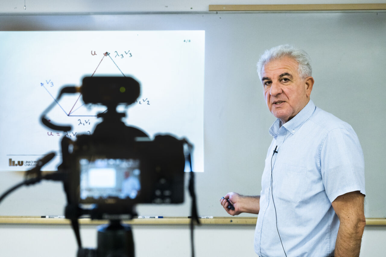 Porträtt av George Baravdish som föreläser framför en whiteboard. Oskarp i förgrunden syns en kamera som spelar in föreläsningen.