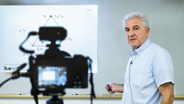 Porträtt av George Baravdish som föreläser framför en whiteboard. Oskarp i förgrunden syns en kamera som spelar in föreläsningen.