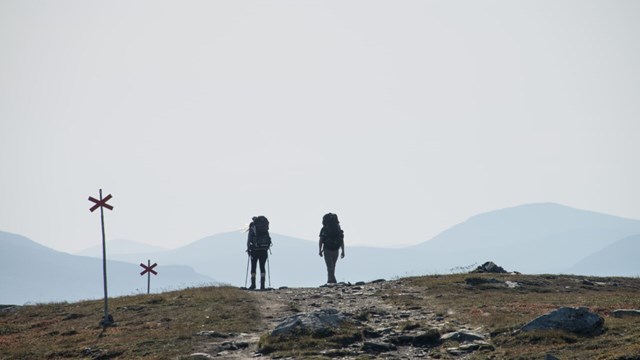Två personer vandrar längs en led på ett berg. Båda har stora ryggsäckar på sig och de går bort från kameran.